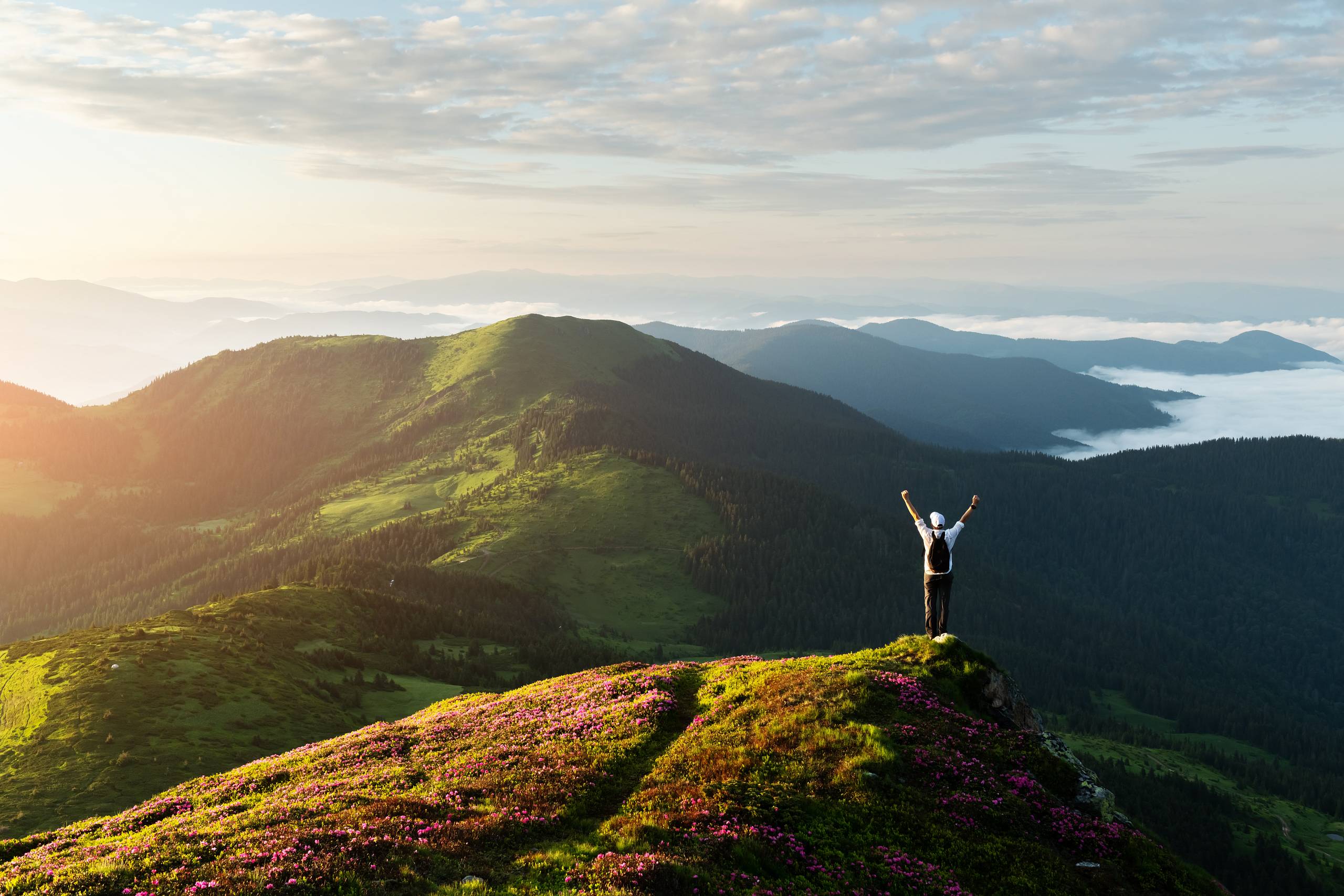 Person mit erhobenen Armen auf Berggipfel bei Sonnenaufgang mit Blick über grüne Berglandschaft.